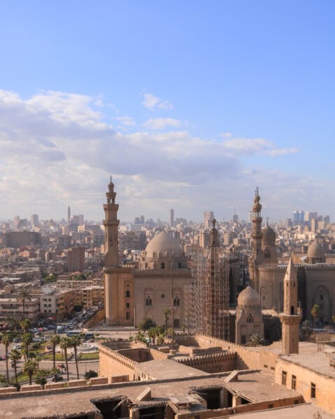 View of a beige coloured stone mosque with two minarets and a domed roof. Behind is a view of a dense city skyline in Cairo Egypt.