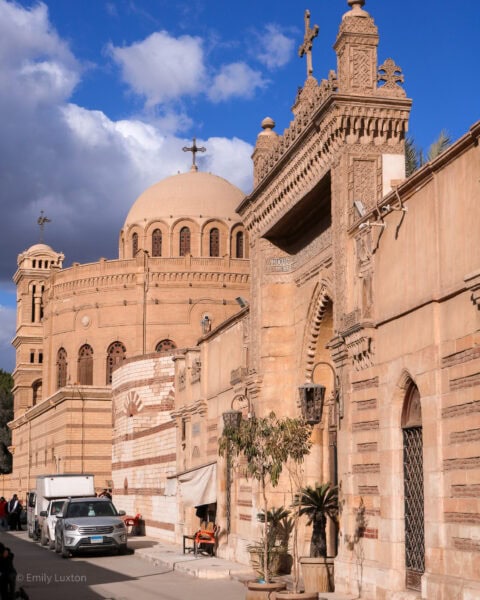 Historic city wall built from beige sandstone with a tall gate. There is a large circular building behind with a domed roof.