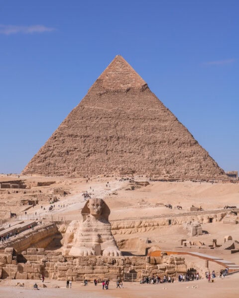 This image depicts two iconic structures of ancient Egypt: the Great Sphinx and the Great Pyramid of Giza. The limestone Sphinx, with the body of a lion and the head of a pharaoh, sits prominently in the foreground, gazing forward. Behind it towers the massive Pyramid of Khafre, the second-largest of the pyramids in Giza, with a smooth, slightly weathered surface against the backdrop of a clear, bright blue sky. Several tourists can be seen walking around the site, adding a sense of scale to the grandeur of these ancient monuments.