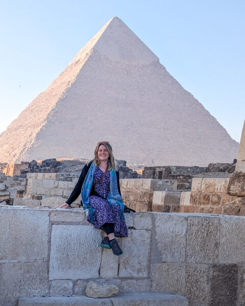 Travel blogger Emily Luxton sitting on a stone wall in front of one of the Great Pyramids at Giza. Emily is wearing a long blue maxi dress with white flowers and a black cardigan with grey trainers and a green scarf. It is just after sunrise and most of the pyramid is in shadow with pale blue sky behind | Solo female travel in Egypt