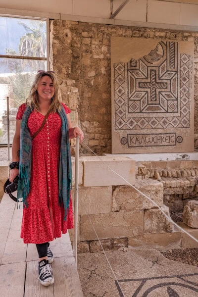 Travel blogger Emily Luxton standing on a wooden bridge next to an excavated ruin with mosaic tiles on the floor below her and on the wall behind. Emily is wearing a long red maxi dress with white flowers, grey trainers and a green scarf.