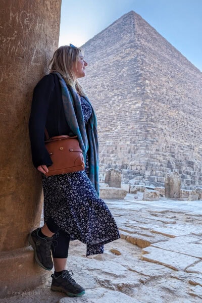 Travel blogger Emily Luxton leaning against an ancient stone pillar looking over her shoulder towards one of the ancient pyramids of Giza. Emily is wearing a long blue maxi dress with white flowers and a black cardigan with grey trainers and a green scarf. 