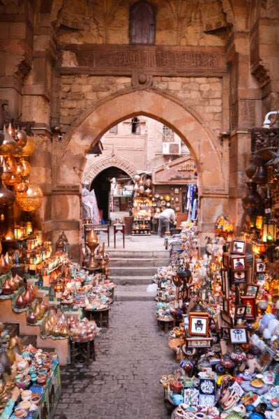 This image showcases a vibrant, bustling market or souk located within an ancient, intricately designed stone archway. The narrow passage is filled with colorful stalls selling an array of traditional goods such as lamps, ceramics, trinkets, and souvenirs. Brass lanterns, pottery, and artwork are neatly arranged, creating a lively and inviting atmosphere. The warm glow from the lanterns adds to the rich ambiance. In the background, additional shops and arches are visible, with a person tending to one of the displays. The historical architecture and the lively market scene suggest a traditional Middle Eastern or North African setting.