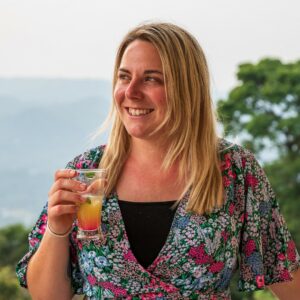 Close up of UK solo female travel blogger Emily wearing a green and pink dress with a floral pattern and holding a glass of an orange coloured cocktail. she is smiling and looking away from the camera and there is a view of hills and jungle trees out of focus behind her. 