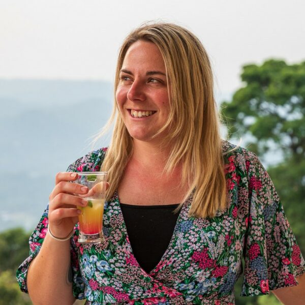 British travel blogger Emily Luxton wearing a floral pink and gren dress and holding a glass with an orange cocktail in, looking away from the camera with a view of trees and hills behind