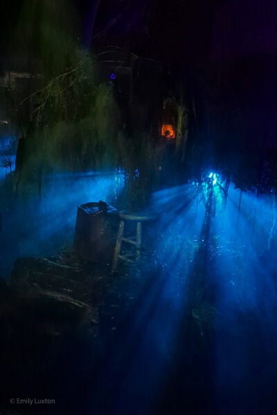 stage set of a swamp with light bursting through a tree and catching on thick fog. It is a very dark night and only the light and fog are visible, as well as a barrel and a stool. Scary theming at Universal Orlando Halloween Horror Nights