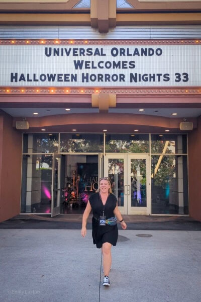 emily wearing a short black dress and a bumbag walking towards the camera in front of a theatre entrance. The sign above the door reads Universal Orlando Welcomes Halloween Horror Nights 33.