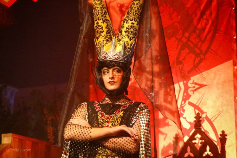 actor in costume as a medieval queen with short black hair wearing a crown with two tall cones made of yellow fabric covered in black lace. she has her arms crossed and is looking off camera. The background behind is red with an image of a face on it.