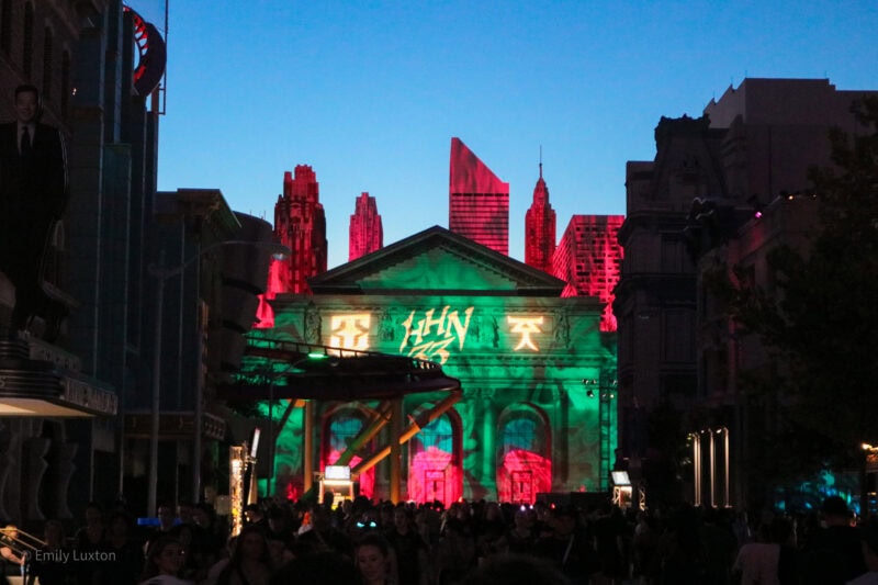 facade of a neoclassical building at night lit in green with a 2D fake city skyline behind it, lit in pink against a clear blue night sky. The letters HHN33 are projected in yellow onto the roof of the building. 

How Scary are Universal Orlando Halloween Horror Nights?