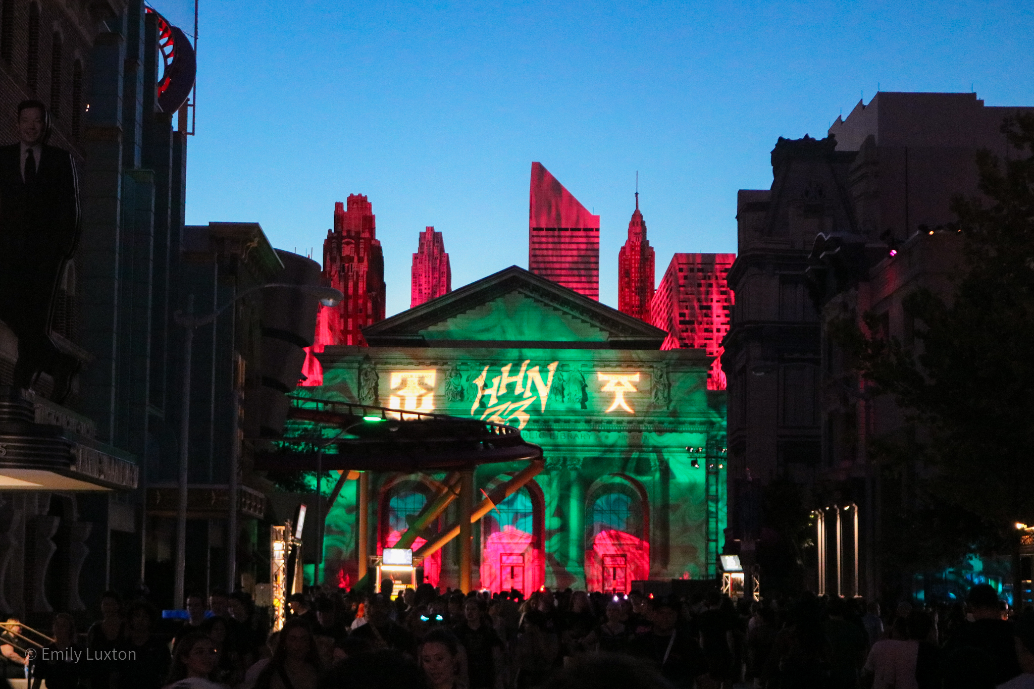 facade of a neoclassical building at night lit in green with a 2D fake city skyline behind it, lit in pink against a clear blue night sky. The letters HHN33 are projected in yellow onto the roof of the building.