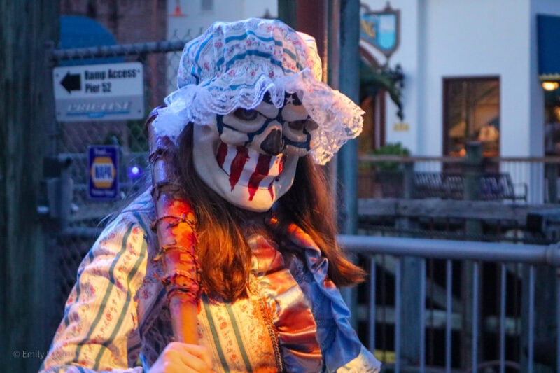 close up of an actor wearing a white clown style mask with blue aroudn the eyes and big red streaks across the grinning mouth. She has long brown hair and a white frilly bonnet on, and is holding a bloody baseball bat over one shoulder.