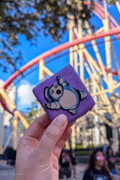 hand holding a small square cake with purple icing and a cartoon of the Ghostbusters Stay Puft Marshmallow man printed on the top, in front of a yellow and red rolldercoaster.