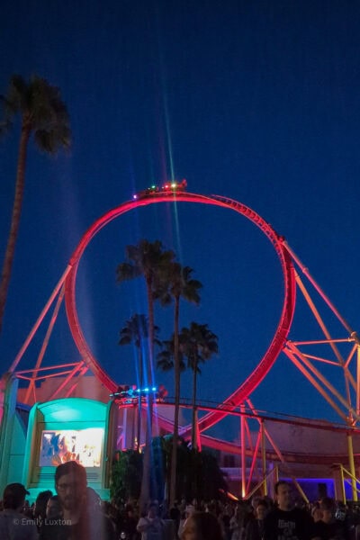 loop the loop on a red rollercoaster at night against a clear dark sky, surrounded by silhouetted palm trees with a crowd of people in the foreground. 