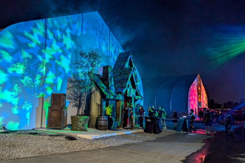 two large white barn style buildings at night lit up, the closest building is lit with green projections and the further one is lit in neon pink. 