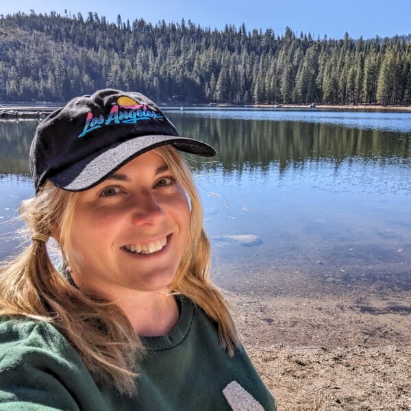 British travel blogger Emily Luxton taking a selfie with a blue lake behind her with a dense forest of pine trees on the far side. She is wearing a dark green sweatshirt and a black baseball cap with Los Angeles written on the front, and her long blonde hair is in loose bunches. 