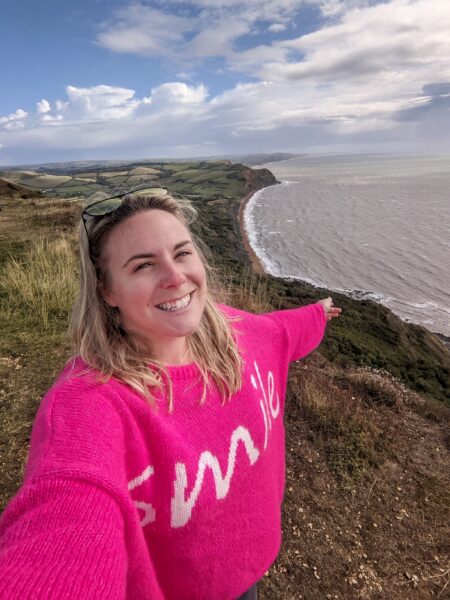 emily wearing a hot pink wooly jumper taking a selfie on top of a cliff with the sea behind her