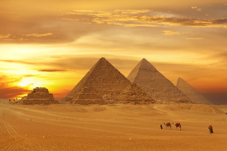 The three Great Pyramids of Giza in a row at sunset with golden sky behind them. There is empty desert in front with the distant figure of a man in long black robes leading three camels in a line across the sand | Solo Female Travel in Egypt tips & advice