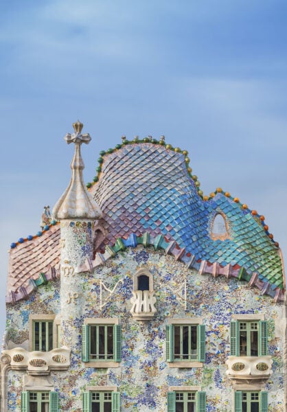 top half of Casa Batllo, a tall building of grey and green stone tiles with a mini turret. The roof is covered with irridescent purple and green tiles that reflect the sun. The sky behind is clear and blue on a sunny day in barcelona. 