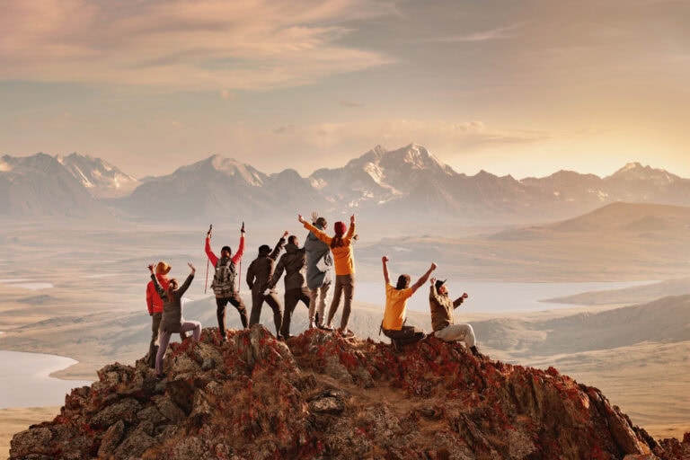 Large group of adults wearing hiking great and raincoats, standing on a rocky mountain peak with a view of distant snowcapped mountains across the valley in front of them at sunrise. They have their arms raised and are facing away from the camera | Best group tours for solo remale travellers