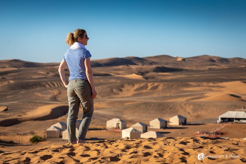 Travel writer Bella standing on a sand dune in a desert looking out towards the sandy desert landsape. There is a small campsite with several cream coloured safari style tents below her. Bella is wearing a blue stripey t-shirt and long khaki trousers and her blonde curly hair is in a ponytail. 