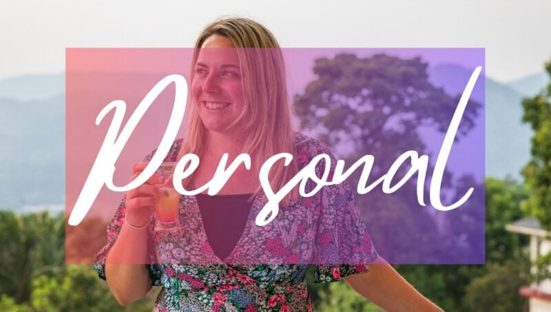 Personal written in white text over an opaque pink box on top of a photo. The photo is a close up of Emily wearing a green and pink dress with a floral pattern and holding a glass of an orange coloured cocktail. she is smiling and looking away from the camera and there is a view of hills and jungle trees out of focus behind her.