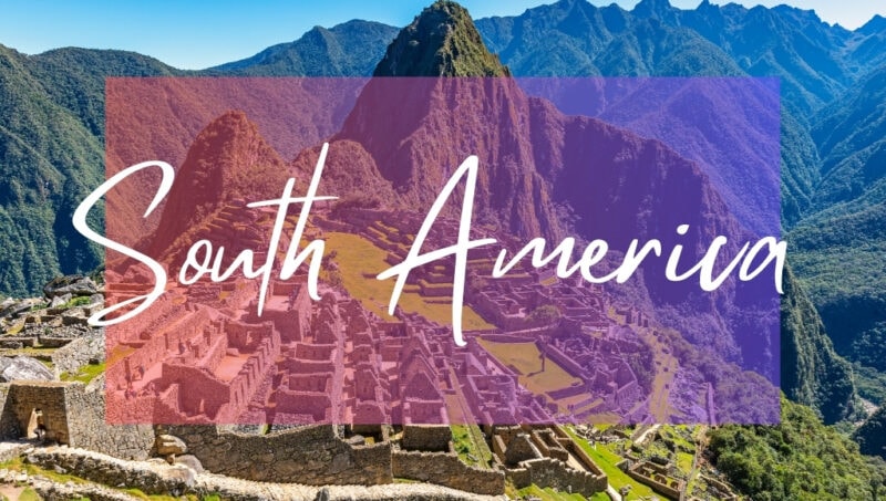 South America written in white text over an opaque pink box on top of a photo of Machu Picchu on a sunny day. There are grey stone Mayan ruins on the grassy slopes of a hill with towering mountain peaks behind and blue sky above.