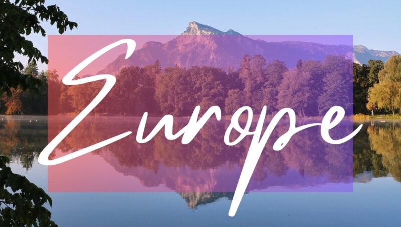 Europe written in white text over an opaque pink box on top of a photo of a mountain surrounded by forest perfectly reflected in a large blue lake with very still water. It is a very sunny day with clear blue sky overhead.