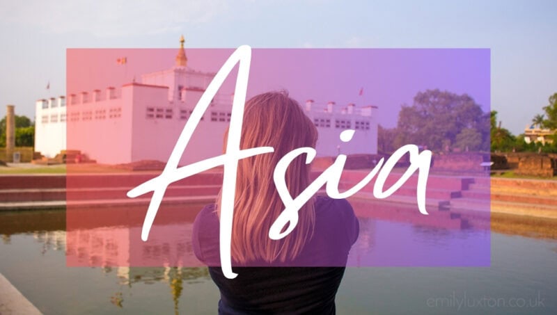 Asia written in white text over an opaque pink box on top of a photo of the back of a woman's head with long straight blonde hair. She is wearing a black t-shirt and facing a pool of blue water with a white temple on the far side on a sunny day in Lumpini Nepal with blue sky above.