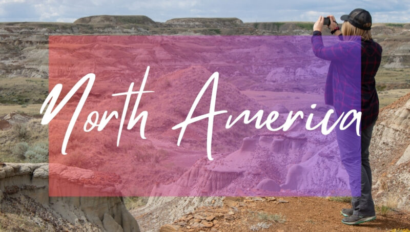 North America written in white text over an opaque pink box on top of a photo of an arid landscape with a rocky valley and beige coloured sandy hills. Travel blogger Emily Luxton is standing on a cliff edge in the foreground holding her camera up to take a photo. She is wearing a dark red checked flannel shirt and grey trousers. 