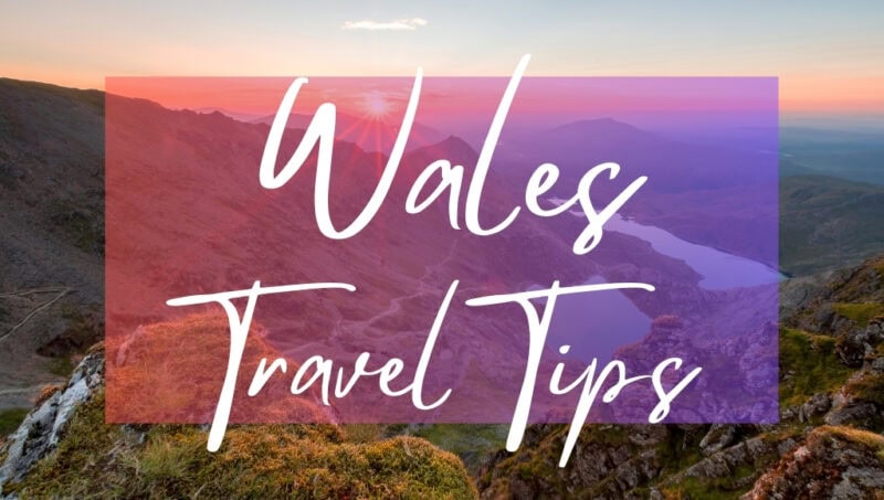 Wales Travel Tips written in white text over an opaque pink box on top of a photo of a lush green landscape in Snowdonia. There are two very blue lakes in the valley below and grassy mountain slopes in the foreground, with the sun just about to set beyond the horizon and pink sky overhead. 