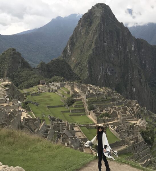 Travel blogger Carina Otero standing on a hilltop in front of a view of Machu Picchu in Peru. There are grey stone walls and ruins of the Incan city spread over a grassy hilltop with a very tall mountain peak behind. Carina is wearing a black t-shirt and black leggings with a long white poncho with black embroidery around the bottom edge, and a large-rimmed straw hat. She is holding her arms out to the sides and smiling at the camera. 