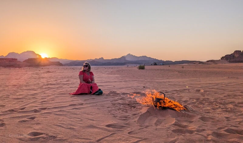 empty desert landscape with the sun setting behind low mountains in the distance. There is a campfire with an iron kettle in the flames and Emily is sat in the sand behind it wearing a red dress. 