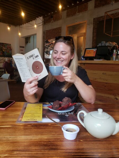 Travel blogger Emily Luxton wearing a black t-shirt sitting alone at a large wooden table in a cafe holding a book in one hand and a grey cup of tea in the other. There is a plate in front of her with a chocolate brownie on it next to a white tea pot and a bowl of sugar. 