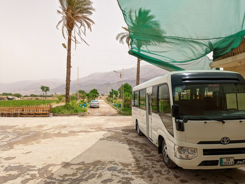 white single decker tour bus parked in a concrete car park under a green awning with two palm trees behind and a road leading away towards a dry mountainous landscape