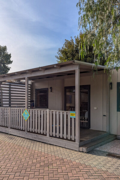 small recangular cabin built from grey wood with a veranda out front surrounded by a grey fence. There are trees on either side and clear blue sky above. Hu Stay Premium mobile home. 