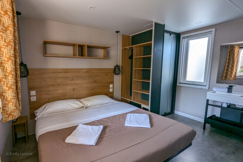 double bed with brown bedspread and white pillows and a large wooden headboard in the bedroom of a mobile home unit. The walls and floor are grey and there are mustard-yellow curtains to the left. Next to the bed is a large shelving unit and on the far right is a stand with a white sink with a mirror above. Hu Stay Premium at Hu Roma Camping in Town. 