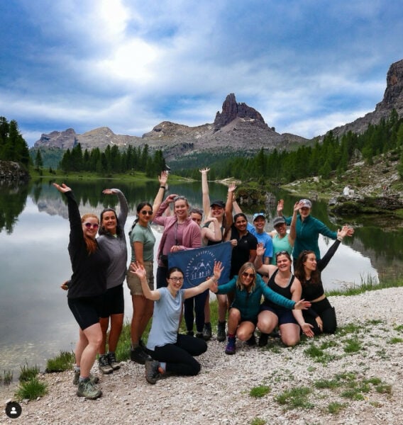 A group of 14 adult women in their 20s and 30s standing in front of a lake with their arms in the air. Two of the girls in the middle are holding a dark blue flag with a picture of camping lodges on it. Behind the lake are rocky mountains adn a dark green forest of evergreen trees | Best group tours for solo female travellers - Outdoor Adventure Girls