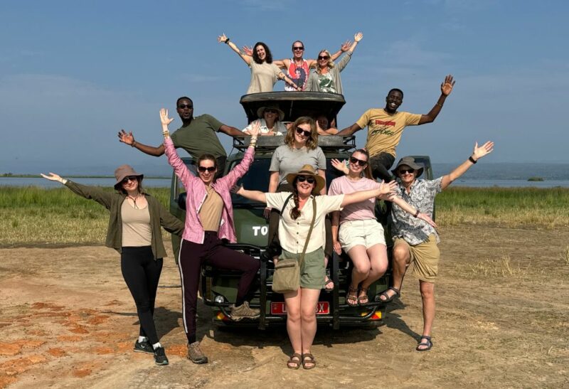 A group of adults standing in front of and on top of a dark green safari jeep in Africa with their arms in the air. The jeep is parked on dry earth with grass and a river behind | Best group tours for solo female travellers - Rock My Adventure group tours in Africa