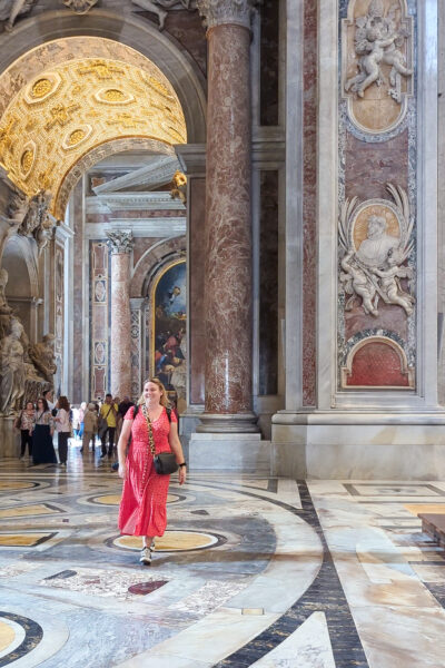 Solo female travel blogger Emily Luxton wearing a long red dress with a ruched waist and small white dots and carrying a small black cross body bag. She is walking towards the camera inside a cathedral with marbel floor and a large marble archway behind her with dark pink pillars and a yellow tiled roof.