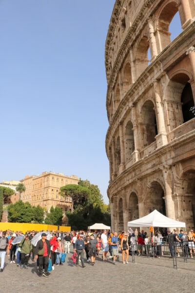 side of the colosseum in rome, a large beige stone circular building with rows of archways around the edge. On the ground in front of the colosseum is a large crowd of people. it is a very sunny day with clear blue sky above.