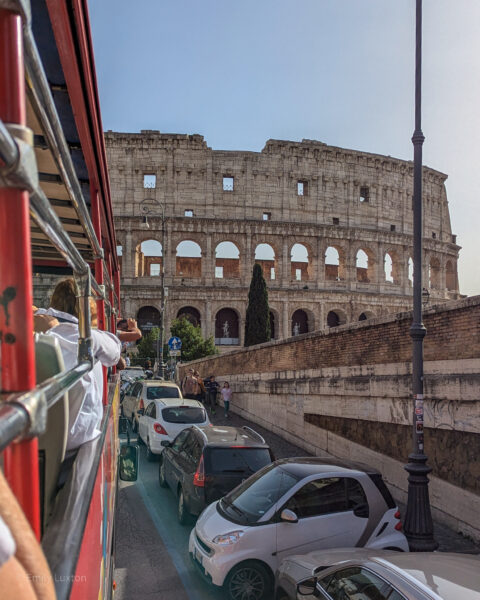 The Colosseum in Rome taken whilst leaning out of the top deck of a double decker bus, the side of the red bus is visible on the left of the photo with parked cars along the road to the right. 