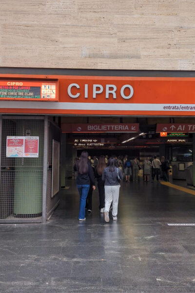 Entry to a metro station in Rome with a black marble floor and grey marble tiled wall. There is a bright orange sign with the name of the station in white text: Cipro