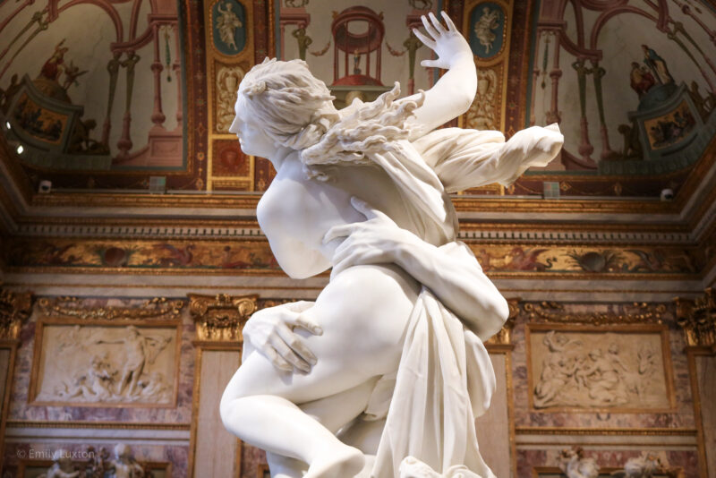 white marble statue depicting a nude woman being grabbed around the waist by a man's arms, she has one arm raised up and her long hair is flying out behind her. Inside the Borghese Gallery in Rome, with the wall behind the statue covered in gold details and engravings. 