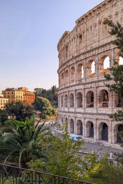 side of the colosseum in rome, a large beige stone circular building with rows of archways around the edge. In the foreground are green leafy trees and a palm tree, with clear blue sky overhead. 