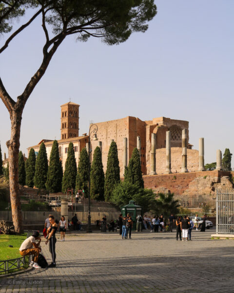 wide pedestrian street wiht grey cobbles where a few people are walking about. The street leads towards a distant Roman ruin where some grey stone pillars and a red stone tower can be seen behind a row of cypress trees. 