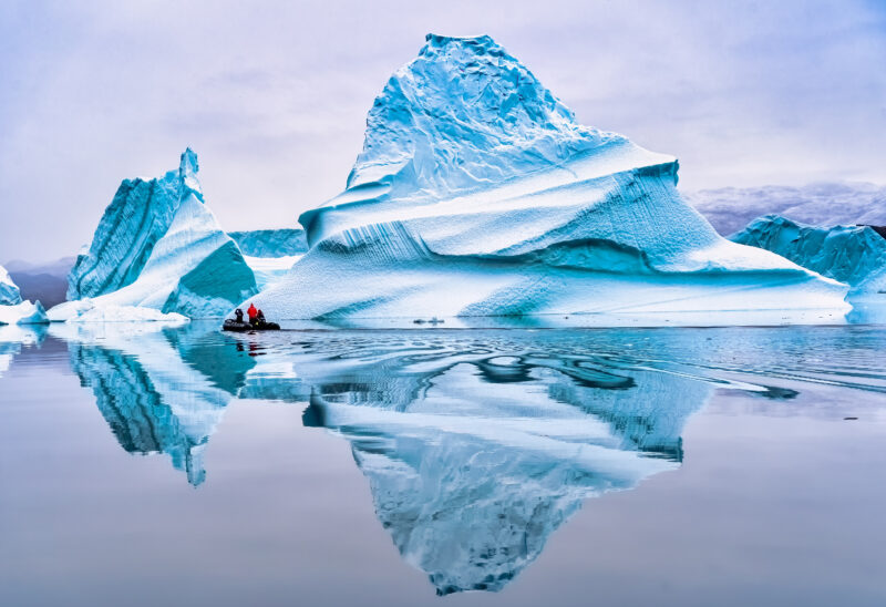 Large white iceberg perfectly reflected in very still water with grey sky behind. There is a small boat with a few human figures alongside the iceberg. 