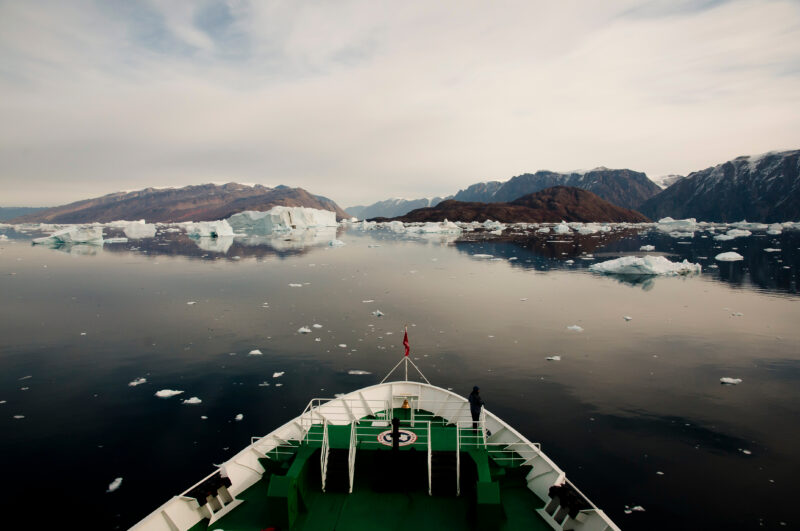 prow of a ship with green deck and white edging surrounded by very calm still grey sea dotted with white iceberds, with several small islands in the distance. 