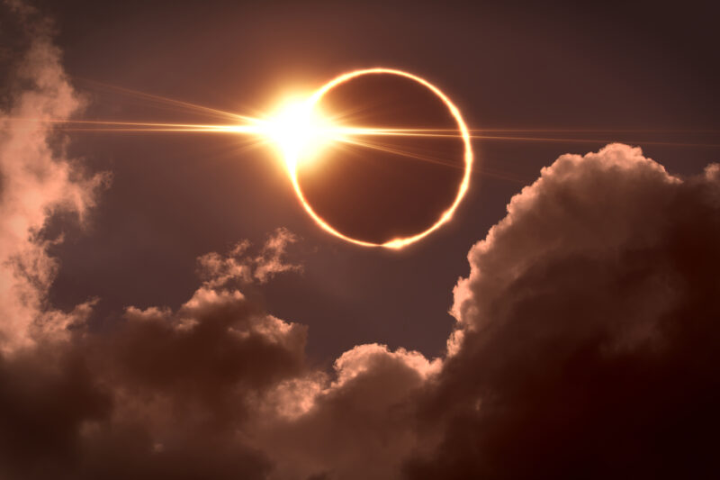 dark sky with a total solar eclipse surrounded by clouds. There is a ring of sunlight and a patch of light flare around the dark circle of the moon