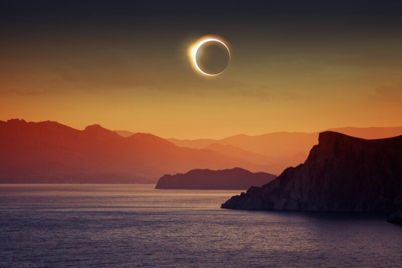 solar eclipse with the moon covering the sun and a ring of light visible around it in the sky above some silhouetted islands with blue sea in the foreground