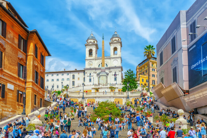 wide stone staircase in Rome with crowds of tourists all over them. There are buildings either side of the steps and a white church at the top with two domed towers and a beige stone obelisk in front. 